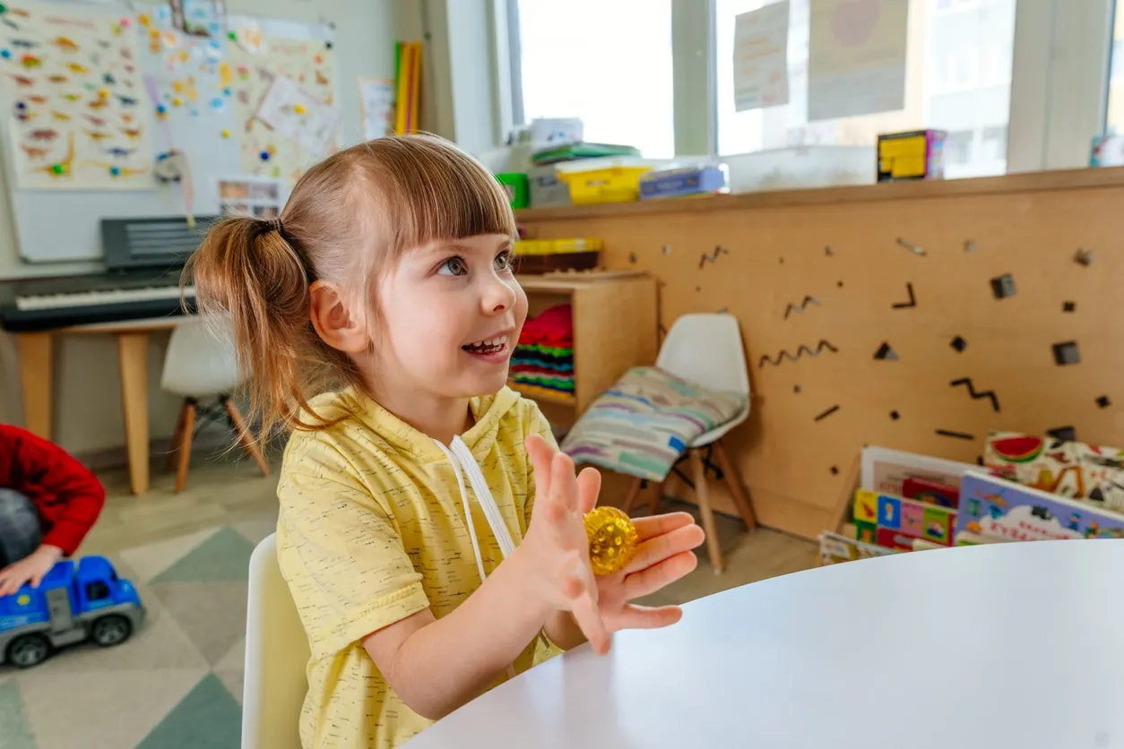 A young girl smiling and exploring a gold sensory ball in a bright, colorful early intervention classroom