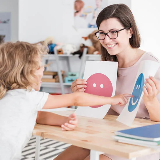 A warm therapist smiling and showing happy and sad emotion cards to a young child during an ABA session