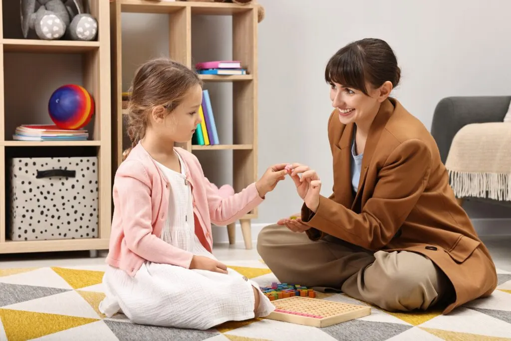 A therapist sitting on the floor playing with wooden blocks with a young girl in a pink cardigan